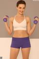 A woman in a white sports bra top and purple shorts holding two purple dumbbells.